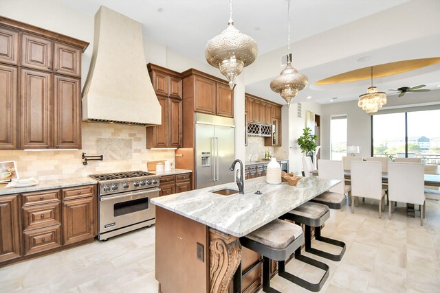 kitchen with decorative backsplash, premium range hood, ceiling fan, high end appliances, and an island with sink