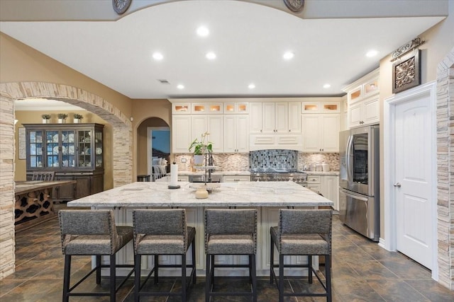 kitchen featuring a large island with sink, decorative backsplash, stainless steel fridge, light stone countertops, and range