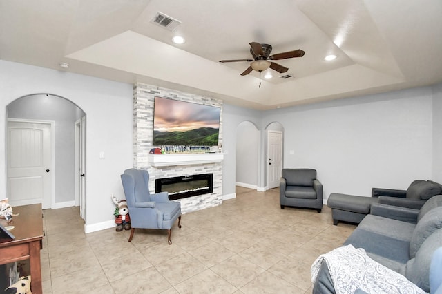 tiled living room with a raised ceiling, a stone fireplace, and ceiling fan