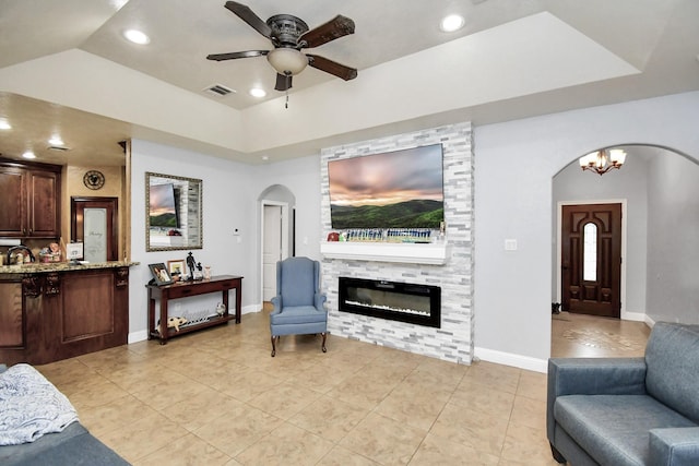 living room with ceiling fan with notable chandelier, a large fireplace, a tray ceiling, and light tile patterned flooring