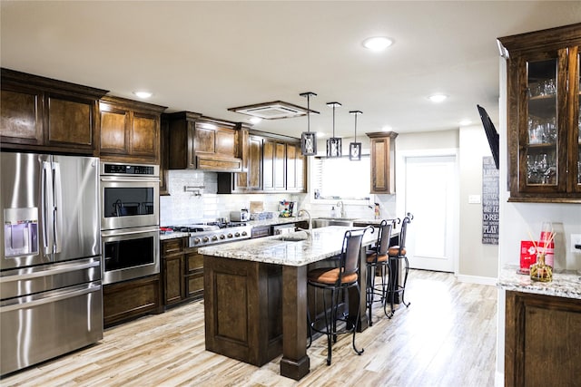 kitchen with a breakfast bar, an island with sink, appliances with stainless steel finishes, decorative light fixtures, and light stone counters