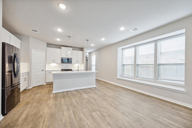 kitchen with appliances with stainless steel finishes, decorative light fixtures, white cabinets, light hardwood / wood-style floors, and an island with sink