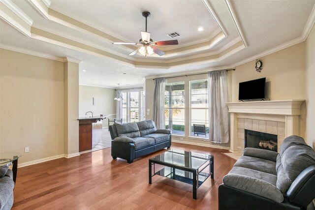 living room featuring ceiling fan, hardwood / wood-style floors, a tray ceiling, a tiled fireplace, and ornamental molding