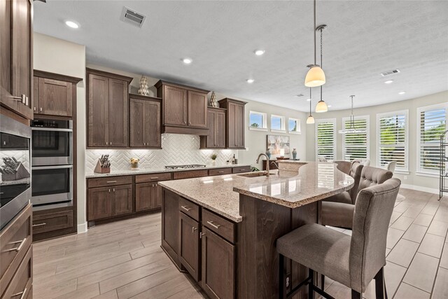 kitchen with a center island with sink, sink, hanging light fixtures, light stone countertops, and a breakfast bar area