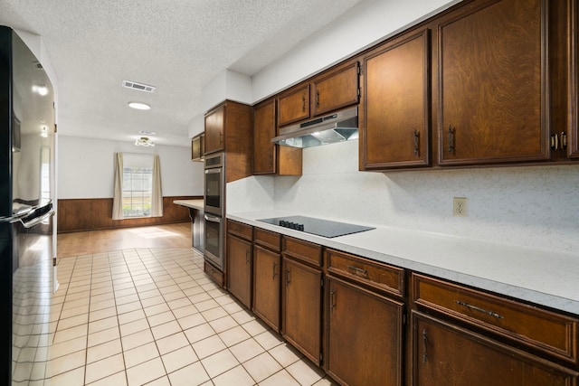 kitchen featuring light tile patterned floors, wooden walls, stainless steel double oven, black electric cooktop, and refrigerator