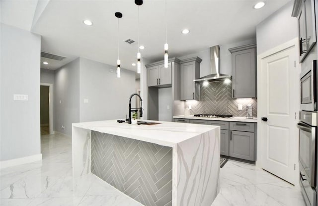 kitchen with a center island with sink, wall chimney range hood, sink, gray cabinets, and decorative light fixtures