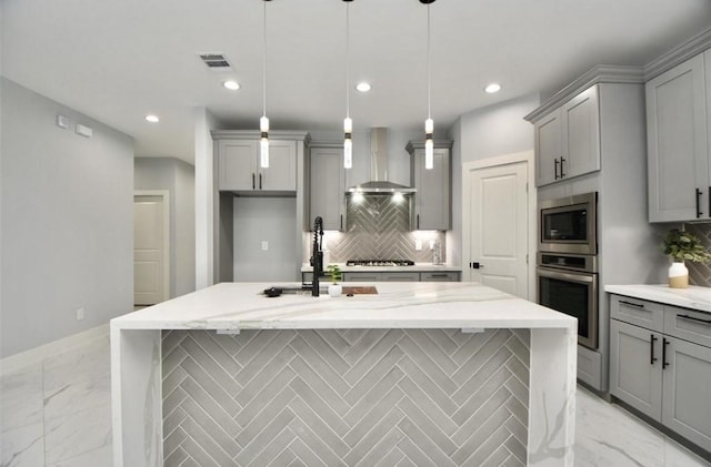 kitchen featuring backsplash, stainless steel appliances, wall chimney range hood, decorative light fixtures, and an island with sink