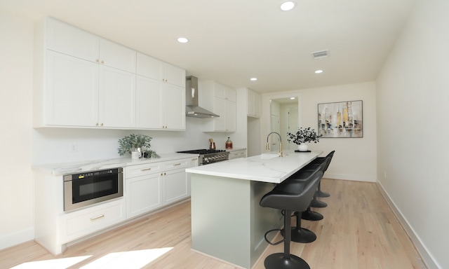 kitchen featuring white cabinetry, wall chimney range hood, a kitchen breakfast bar, a kitchen island with sink, and light wood-type flooring