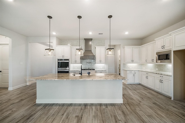 kitchen with premium range hood, a center island with sink, white cabinets, light stone counters, and stainless steel appliances