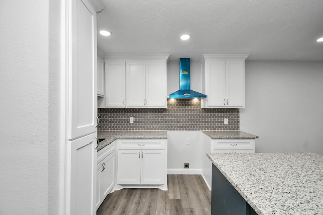 kitchen featuring tasteful backsplash, light stone counters, wall chimney exhaust hood, white cabinets, and light hardwood / wood-style floors