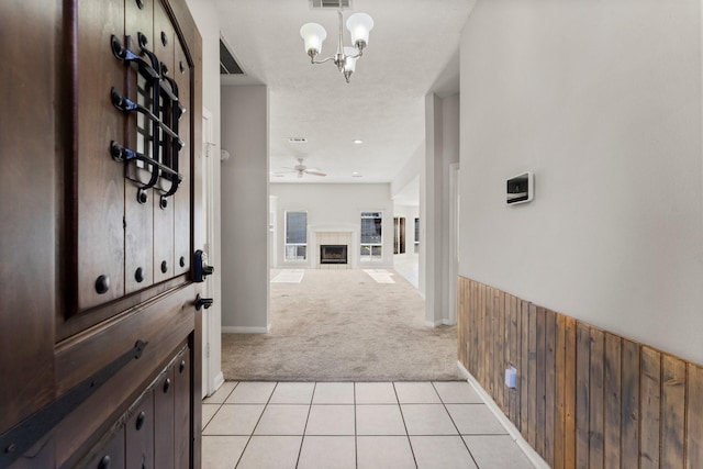 foyer entrance featuring ceiling fan with notable chandelier, light colored carpet, a tiled fireplace, and wood walls