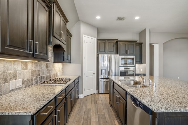 kitchen with appliances with stainless steel finishes, light wood-type flooring, dark brown cabinets, a kitchen island with sink, and sink