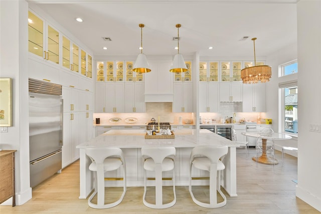 kitchen with built in fridge, light stone countertops, pendant lighting, white cabinets, and a center island with sink