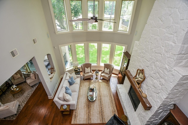 living room with dark hardwood / wood-style flooring, a towering ceiling, a stone fireplace, and ceiling fan