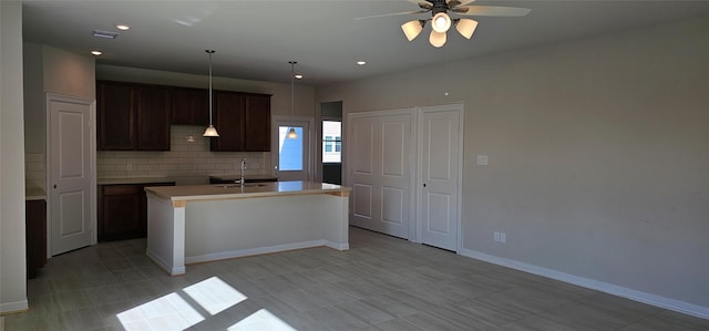 kitchen with backsplash, sink, hanging light fixtures, ceiling fan, and an island with sink