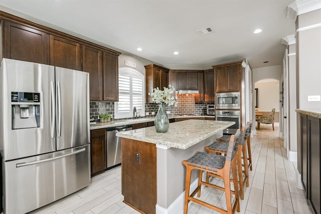 kitchen featuring backsplash, stainless steel appliances, light stone counters, dark brown cabinetry, and a kitchen island