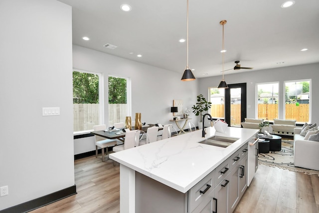 kitchen featuring ceiling fan, sink, hanging light fixtures, light stone counters, and a center island with sink