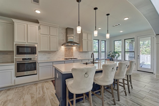 kitchen featuring a kitchen island with sink, stainless steel appliances, pendant lighting, sink, and wall chimney range hood