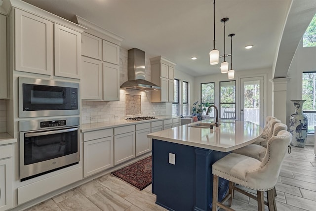 kitchen featuring hanging light fixtures, wall chimney range hood, a kitchen island with sink, appliances with stainless steel finishes, and sink