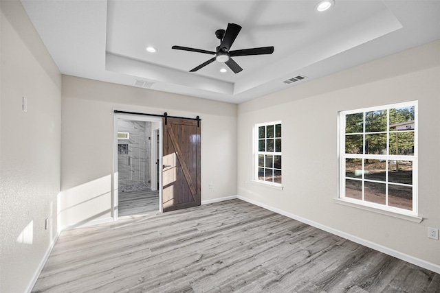 unfurnished room featuring ceiling fan, a barn door, light wood-type flooring, and a tray ceiling