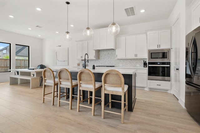 kitchen featuring backsplash, white cabinets, an island with sink, decorative light fixtures, and stainless steel appliances