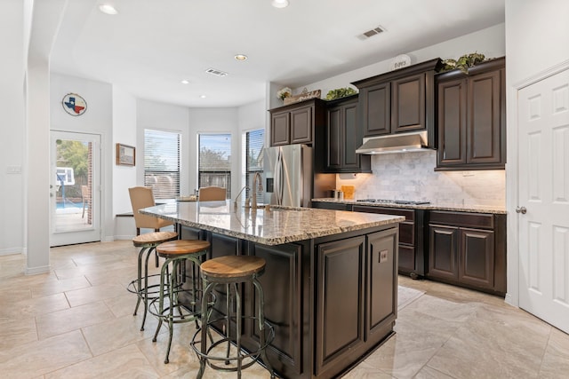 kitchen featuring dark brown cabinetry, light stone countertops, stainless steel appliances, a kitchen bar, and a center island with sink