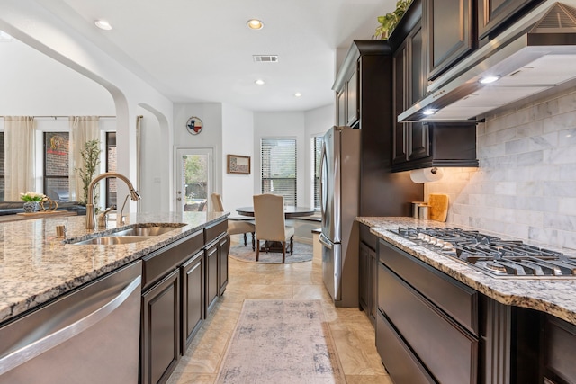 kitchen with appliances with stainless steel finishes, backsplash, light stone counters, sink, and range hood