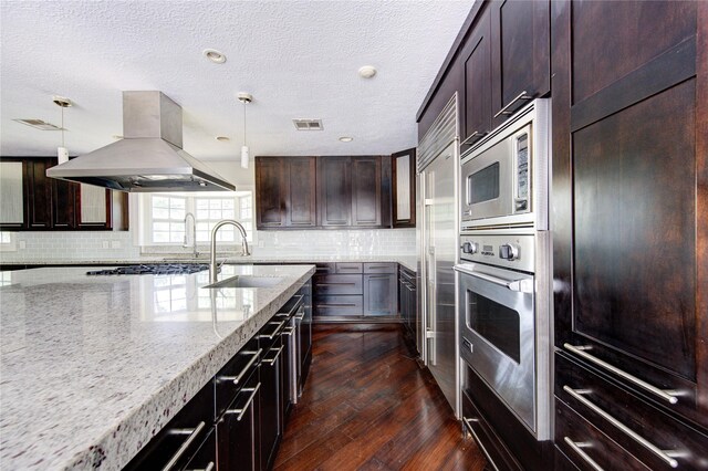 kitchen with light stone countertops, sink, island exhaust hood, built in appliances, and decorative light fixtures