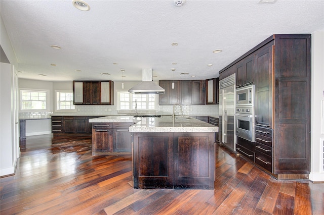 kitchen with dark brown cabinetry, extractor fan, a kitchen island with sink, built in appliances, and decorative light fixtures