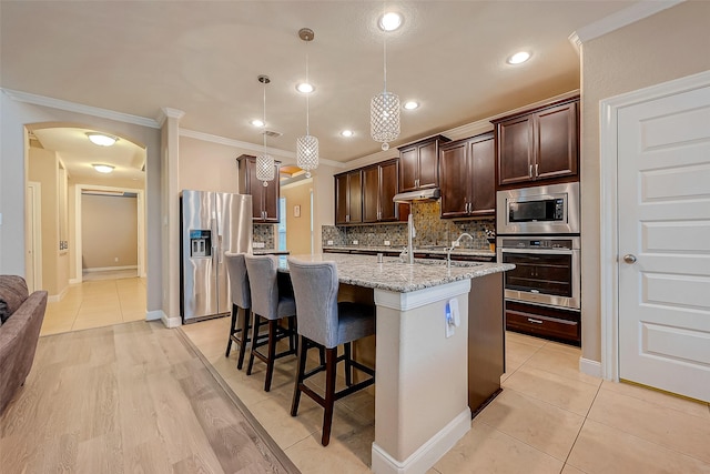 kitchen with decorative backsplash, appliances with stainless steel finishes, light stone counters, a center island with sink, and decorative light fixtures