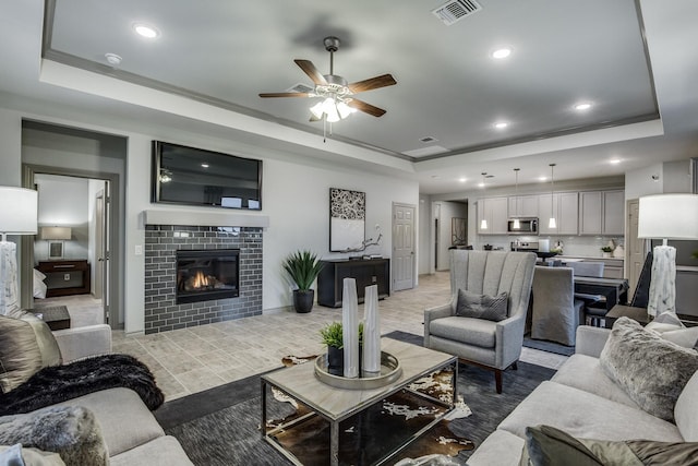 living room featuring ceiling fan, a raised ceiling, ornamental molding, and a tile fireplace