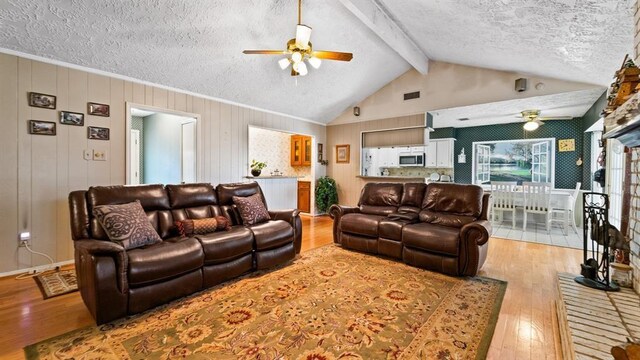 living room with light wood-type flooring, a brick fireplace, a textured ceiling, ceiling fan, and beamed ceiling