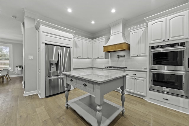 kitchen with stainless steel appliances, white cabinets, light stone counters, premium range hood, and crown molding