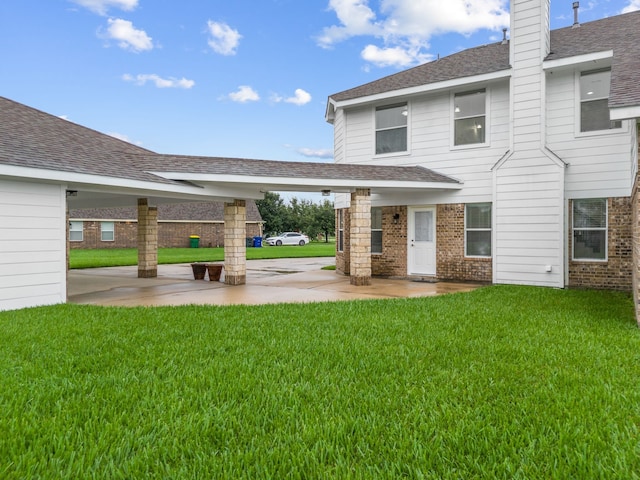 back of property with a shingled roof, brick siding, a patio, and a lawn