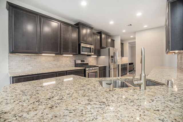 kitchen with stainless steel appliances, backsplash, light stone countertops, dark brown cabinetry, and sink