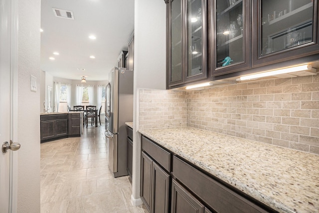 kitchen featuring light stone countertops, stainless steel fridge, dark brown cabinets, and tasteful backsplash