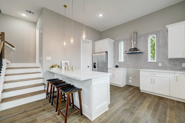 kitchen with wood-type flooring, stainless steel fridge, kitchen peninsula, wall chimney range hood, and white cabinets