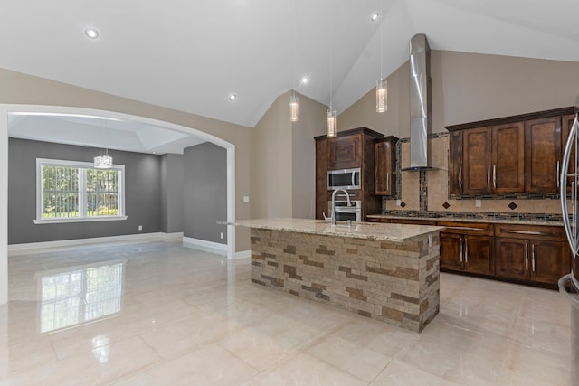 kitchen featuring wall chimney range hood, stainless steel appliances, tasteful backsplash, a kitchen island with sink, and light stone counters