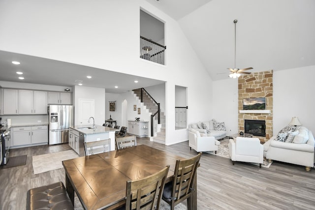 dining space with a stone fireplace, ceiling fan, sink, high vaulted ceiling, and light hardwood / wood-style flooring