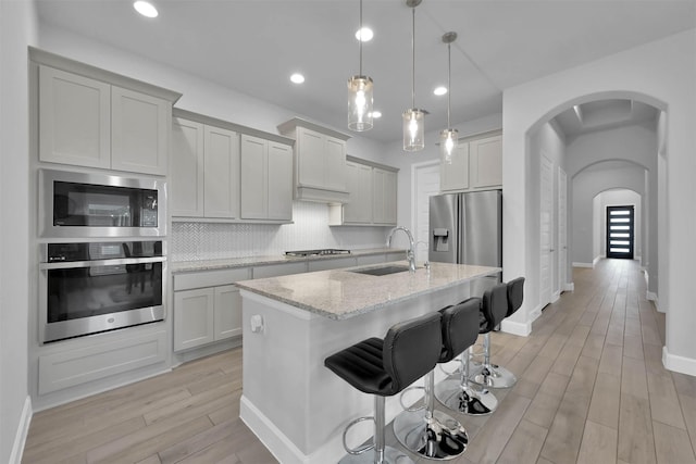 kitchen with sink, decorative backsplash, an island with sink, appliances with stainless steel finishes, and light stone counters