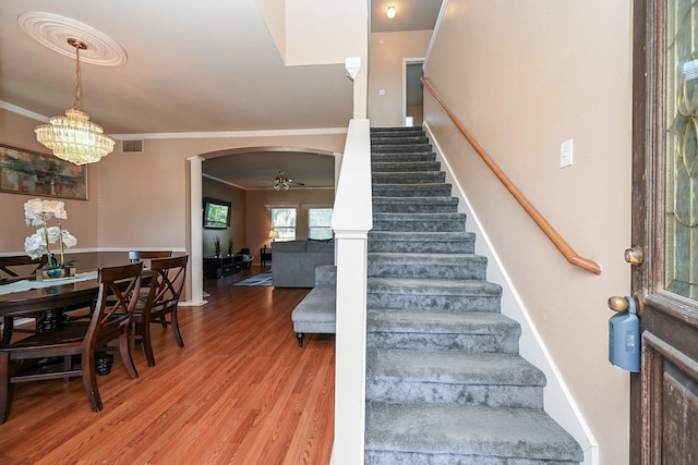 stairs with wood-type flooring, ceiling fan with notable chandelier, and ornamental molding