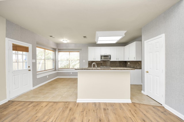 kitchen with tasteful backsplash, dark stone counters, light hardwood / wood-style flooring, white cabinets, and an island with sink