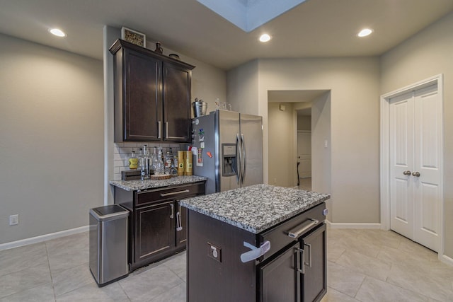 kitchen with stainless steel refrigerator with ice dispenser, tasteful backsplash, light stone counters, dark brown cabinetry, and a kitchen island