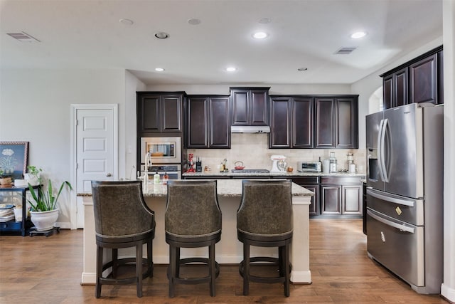 kitchen with light stone countertops, dark hardwood / wood-style flooring, stainless steel appliances, and a kitchen island with sink