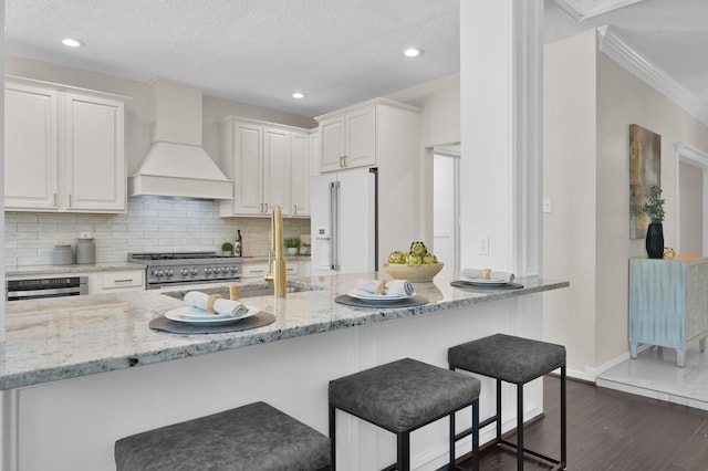 kitchen featuring high end white refrigerator, custom exhaust hood, gas range oven, a kitchen breakfast bar, and white cabinets