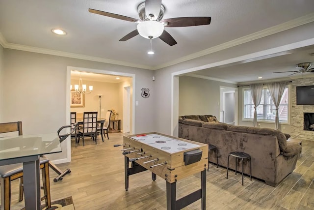 playroom featuring a stone fireplace, crown molding, light hardwood / wood-style floors, and ceiling fan with notable chandelier