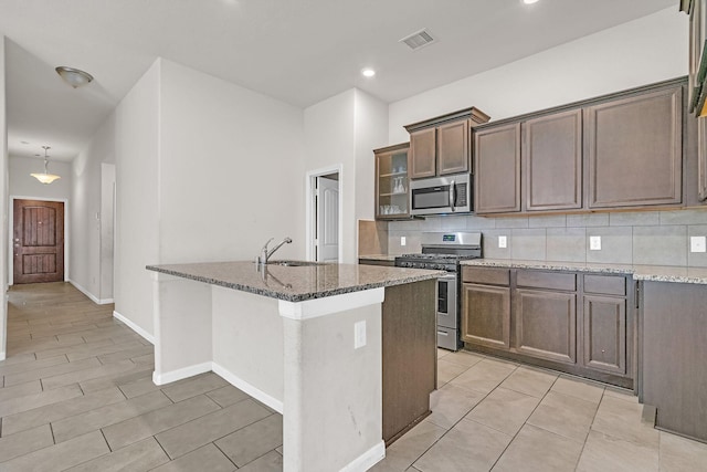 kitchen with backsplash, a kitchen island with sink, dark stone counters, sink, and appliances with stainless steel finishes