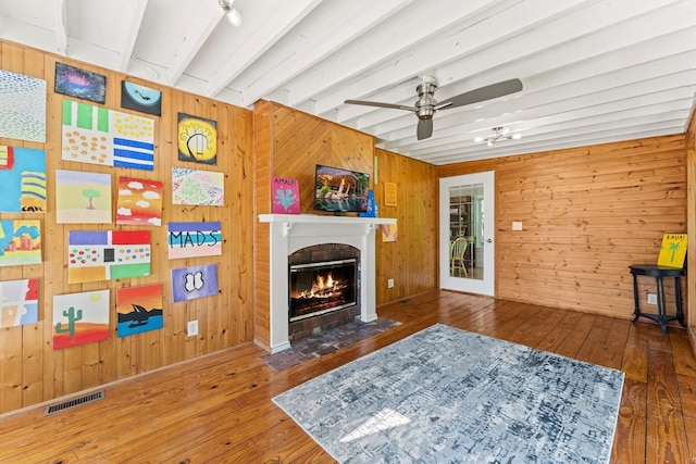 living room featuring a brick fireplace, ceiling fan, beamed ceiling, hardwood / wood-style floors, and wood walls