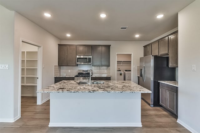 kitchen with light wood-type flooring, dark brown cabinetry, stainless steel appliances, a kitchen island with sink, and washing machine and dryer