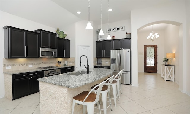 kitchen featuring appliances with stainless steel finishes, hanging light fixtures, an island with sink, light stone countertops, and backsplash